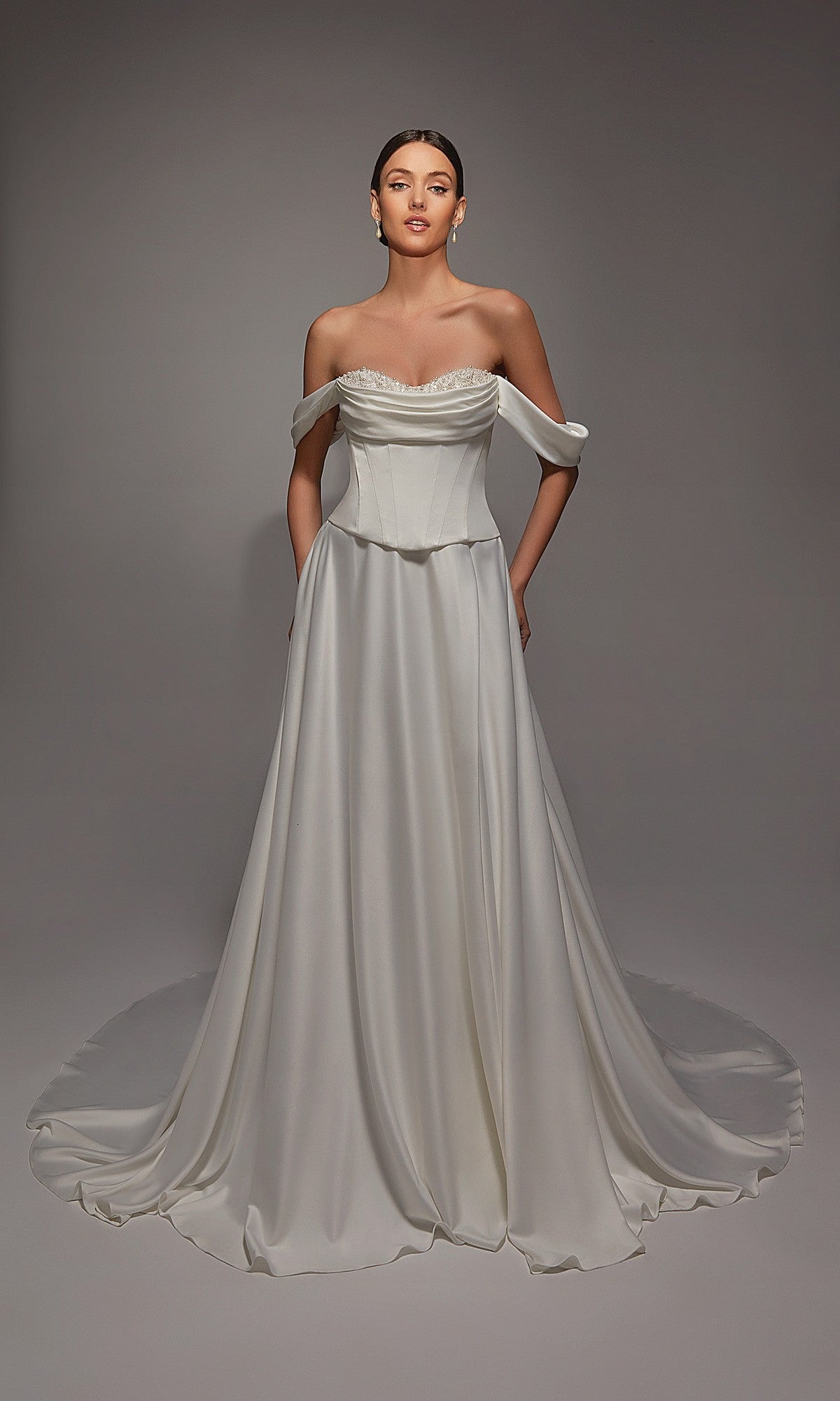 A woman stands in a studio wearing the Alyce 7142 long white dress, featuring an off-the-shoulder satin design with a fitted corset bodice and a floor-length flowing skirt with a train.